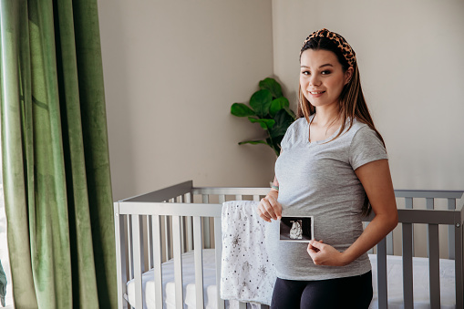 Anticipating the arrival of her new baby, a pregnant mother-to-be cherishes a moment in the nursery while viewing an ultrasound photo.