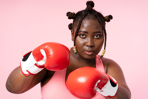 Portrait of beautiful serious Nigerian woman wearing top, boxing gloves, looking at camera, doing punch, breast cancer fight concept, isolated on pink background