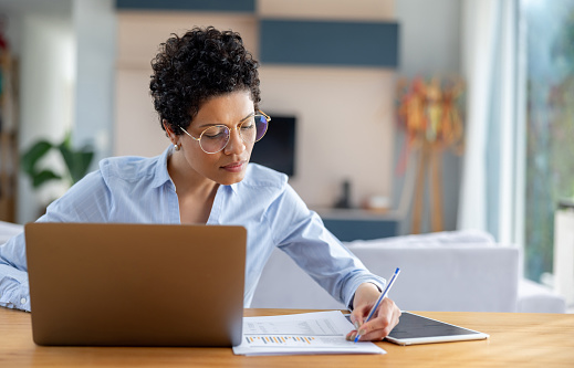 African American businesswoman working at home using her laptop and writing notes - telecommuting concepts