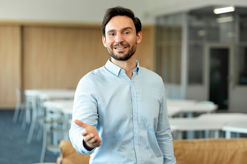 Handsome smiling bearded man working in modern office, attractive businessman with outstretched hand, greeting, gesturing looking at camera. Concept of successful business
