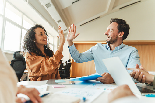 Smiling handsome businessmen, bearded hispanic man and african american woman giving high five to each other sitting in modern office. Corporation concept