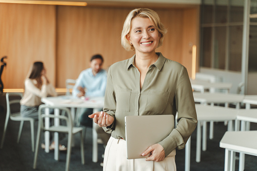 Portrait of smiling middle aged businesswoman holding laptop working in modern office, standing, looking away. Professional saleswoman at meeting with group of businesspeople