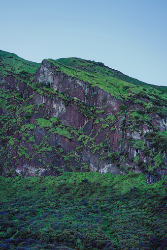 Stunning outdoor mountain landscape with lush green grass, rocky terrain, hillside, cliff face, beautiful blue sky.