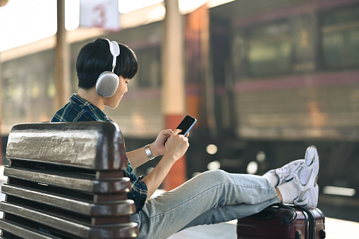 Young man in headphone using mobile phone during waiting for train at railway station.