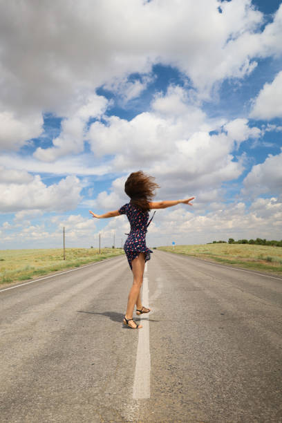 A photo of a barefoot woman in a black dress, with her back on an empty road.
