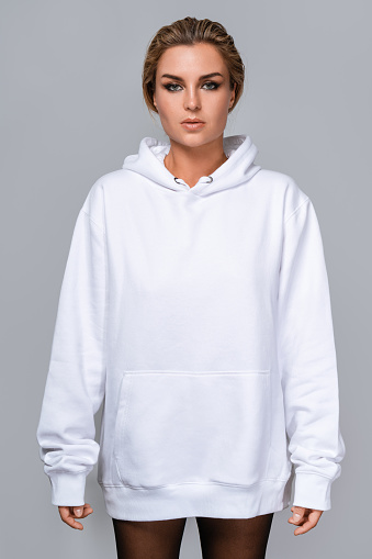 Woman dressed in a white oversized hoodie with blank space, ideal for a mockup, set against gray background.