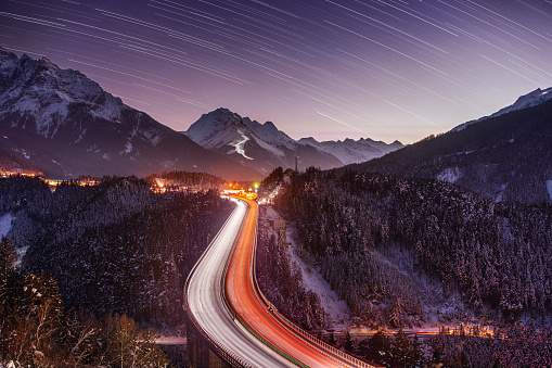 highway bridge with light trails through european alps, mountains in tyrol austria on winter evening with snow and star trails on night sky