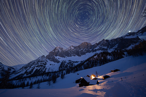 star trail on night sky over mountain range in winter with snow and illuminted wooden huts, cottages in idyllic austrian alps, europe