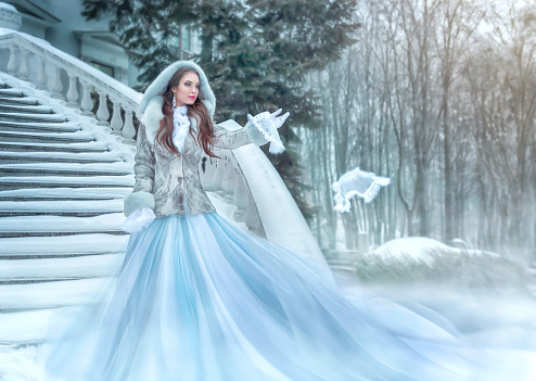 A beautiful young brunette in a long blue dress stands on a white staircase under the snow in winter. Throws down a small white lace handkerchief. artistic portrait