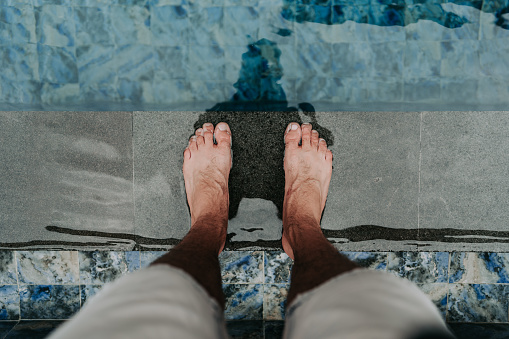 A person's feet stand on the pool edge, surrounded by light blue tiles, suggesting a refreshing dip in an outdoor swimming pool or beach area during hot summer