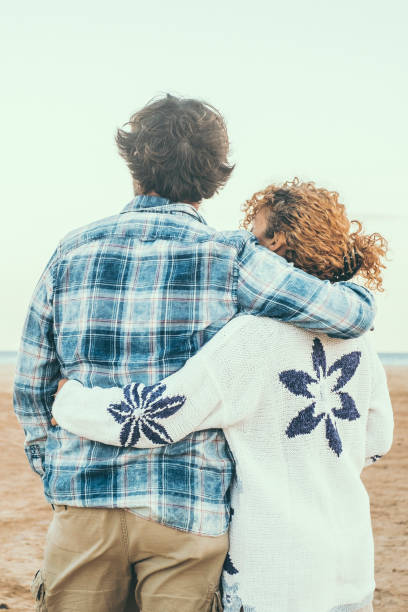 rear view portrait of couple in love admiring the beach and ocean hugging with love. romantic life in winter holiday vacation. people in relationship embracing. love and togetherness concept - honeymoon beach couple heterosexual couple fotografías e imágenes de stock