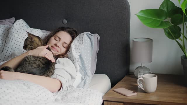 A young Caucasian woman and her gentle gray cat are relaxing lying in bed