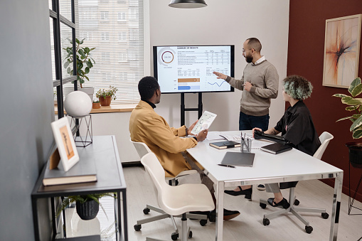 Multi-ethnic group of young coworkers discussing infographics presented on screen in conference room