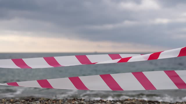 Red white warning tape barrier ribbon swinging in the wind across exotic sea beach background without people. No entry Red White caution tape. No holiday concept, delayed travel, no summer plans