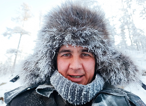 close-up portrait of a smiling Caucasian middle-aged man in a winter fur hat in the forest and with frost on his face from the frost. Winter walk in the forest