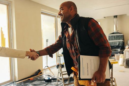 Happy mature contractor shakes hands with a homeowner after signing the paperwork, finalizing a home renovation deal. A successful collaboration for an indoor remodel and improvement project.