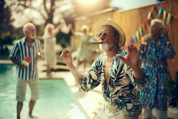 Carefree mature man with a cigar dancing on a pool party with his friends. Young at heart senior man having fun while smoking a cigar and dancing during a pool party in the backyard. His friends are in the background. man cigar stock pictures, royalty-free photos & images