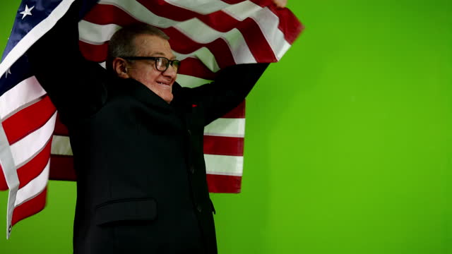 Senior man in black suit and eyeglasses raises American flag as sign of victory.