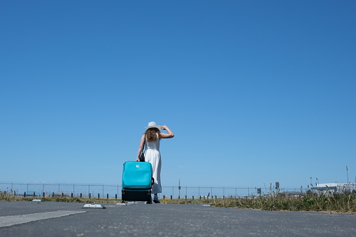 woman girl in white dress tourist with large blue suitcase goes into distance adjusting hat against sky Travel runway place for travel agency advertising text. beautiful developing long clothes