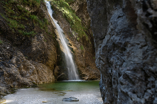 A waterfall with a pool and a stream bed with clear water in the mountains, green trees.