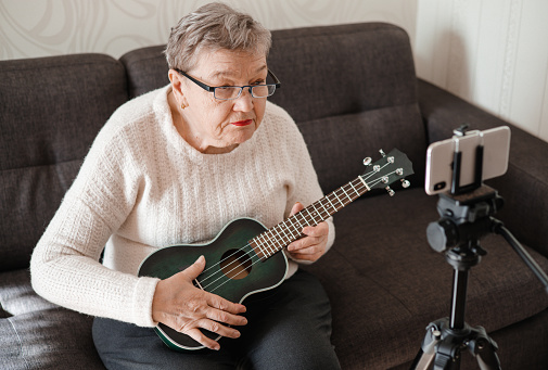 Elderly woman 70+ sits on the couch and learns to play the ukulele at home using video tutorial on her smartphone. Video course on learning to play the guitar. Adaptation of pensioners in the modern world. New knowledge and skills. Prevention of Alzheimer's disease, sclerosis or dementia. Using of modern technologies