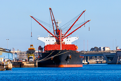 Large bulk carrier cargo ship unloading at port on a sunny day; symmetry with crane boom positions; large orange bucket (with 