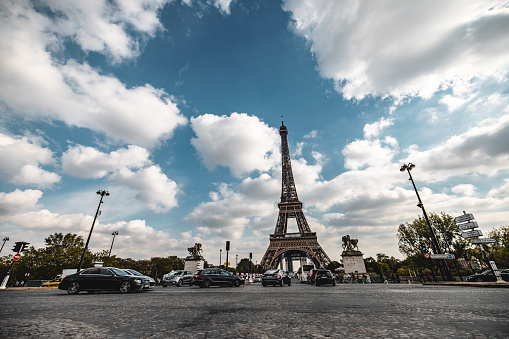 Street level view of the iconic Eiffel Tower in Paris with vehicles and blue cloudy sky.
