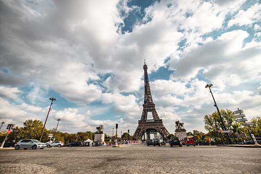 Wide shot of the iconic Eiffel Tower in Paris, France, with tourists and vehicles on a partly cloudy day.