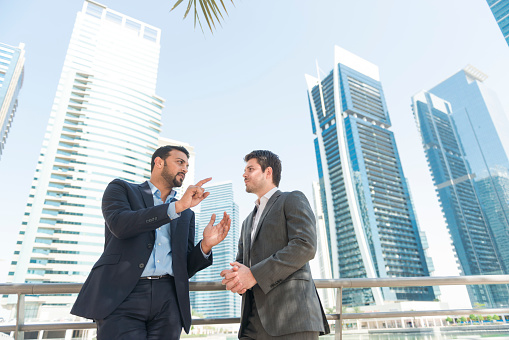 Horizontal color image of one Arabic and one western businessmen talking in front of the Dubai skyline. Copy space above and left.