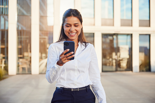 Smiling businesswoman walking outside the office using mobile phone texting message