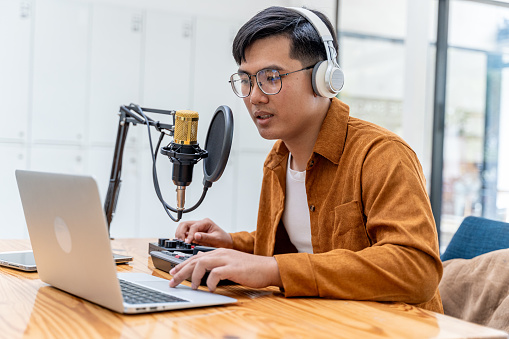 Young Asian man speaking to microphone during his live podcast while checking the feedback from his online audiences from his laptop computer