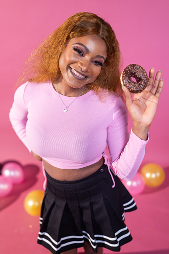 A beaming woman showcases a chocolate donut, her delightful expression matching the playful charm of her pink outfit and the festive atmosphere