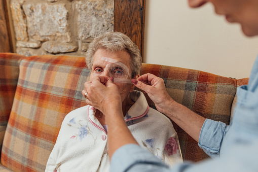 A senior woman and her mature daughter at home in Seghill, Northumberland. The senior woman is staying with her daughter for support and care as she has dementia. They are in the living room in the morning and the daughter is applying an eye patch to her mother's eye because she is recovering from a cataract operation.