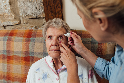 A senior woman and her mature daughter at home in Seghill, Northumberland. The senior woman is staying with her daughter for support and care as she has dementia. They are in the living room in the morning and the daughter is applying eyedrops to her mother's eye because she is recovering from a cataract operation.