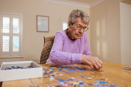 A senior woman at home in Seghill, Northumberland. The senior woman is staying with her daughter for support and care as she has dementia. She is sitting at the dining table by herself, doing a jigsaw puzzle to help with her memory.