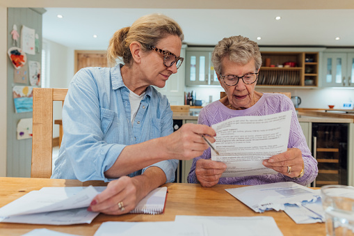A senior woman and her mature daughter at home in Seghill, Northumberland. The senior woman is staying with her daughter for support and care as she has dementia. They are sat at the dining room table where they're looking through financial bills, looking concerned.