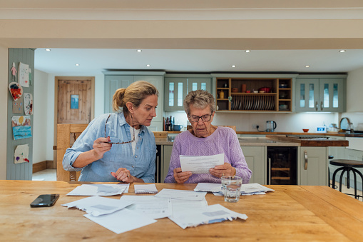 A senior woman and her mature daughter at home in Seghill, Northumberland. The senior woman is staying with her daughter for support and care as she has dementia. They are sat at the dining room table where they're looking through financial bills, looking concerned.