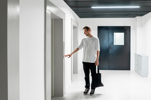 A young man stands poised to press the elevator call button in the sleek hallway of a contemporary building.