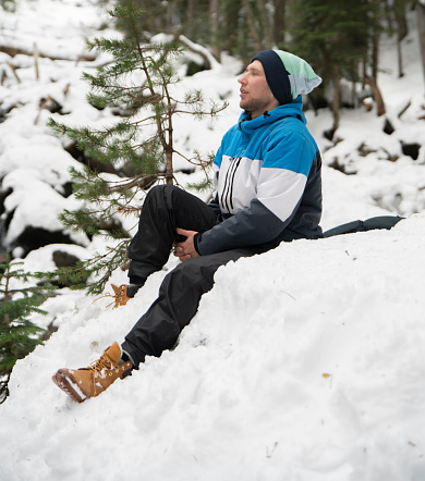 A solitary man sits on a blanket of fresh snow, surrounded by a serene, snow-covered forest, seemingly lost in thought or enjoying the tranquility of winter.