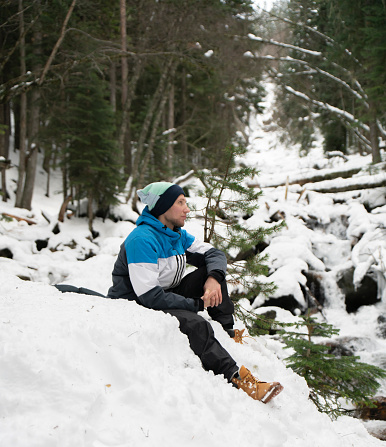 A solitary man sits on a blanket of fresh snow, surrounded by a serene, snow-covered forest, seemingly lost in thought or enjoying the tranquility of winter.