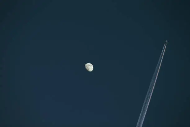 Photo of Photo of moon during day with airplane vapor trail in the sky.
