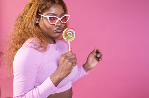 A stylish woman in pink balances a lollipop on her lips, her fashionable sunglasses adding an air of playful mystery.