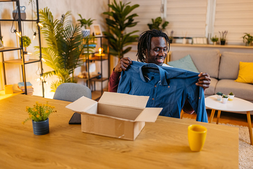 Handsome young black man sitting at the kitchen table and unboxing a package at night with a new button down shirt he ordered online.