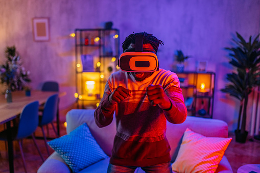 Handsome young black man playing a boxing virtual reality game on a VR headset at home in the living room at night.