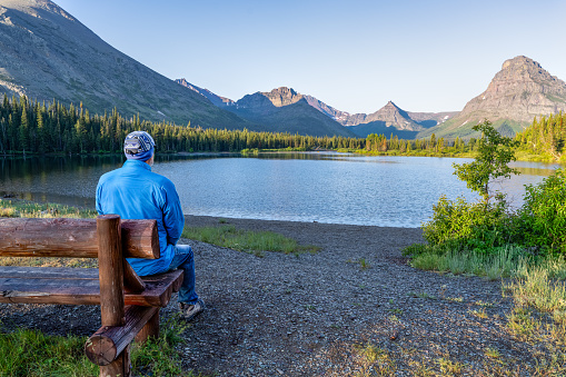 Man, person sitting watching a scene of a lake surrounded by trees and a rugged mountain in the background, Pray Lake, Two Medicine Area, Glacier National Park, Montana
