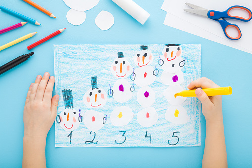 Little child hands creating and decorating snowman shapes and learning numbers on paper on light blue table background. Pastel color. Closeup. Point of view shot. Toddler development. Top down view.