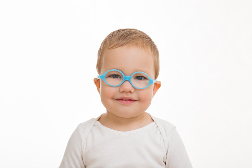 1 year and 8 month old cute smiling baby boy in white clothes with blue eyeglasses isolated on light gray background. Front view. Closeup. Looking at camera.