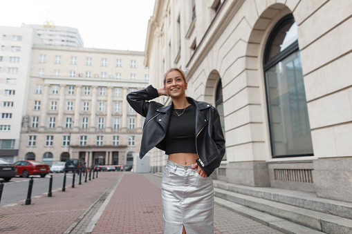 Happy beautiful young woman model with a smile in fashion black clothes with a leather jacket and top and skirt walking in the city. Pretty chic lady