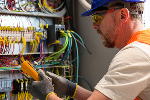 electrician in blue helmet, yellow glasses and orange vest measures electric current with digital multimeter in distribution box-blurred background.