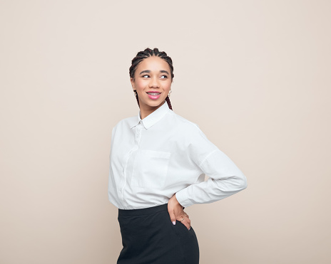 Multiracial teenage girl with braided hair wearing white shirt and black skirt standing with hands on hips and looking away. Studio shot, beige background.